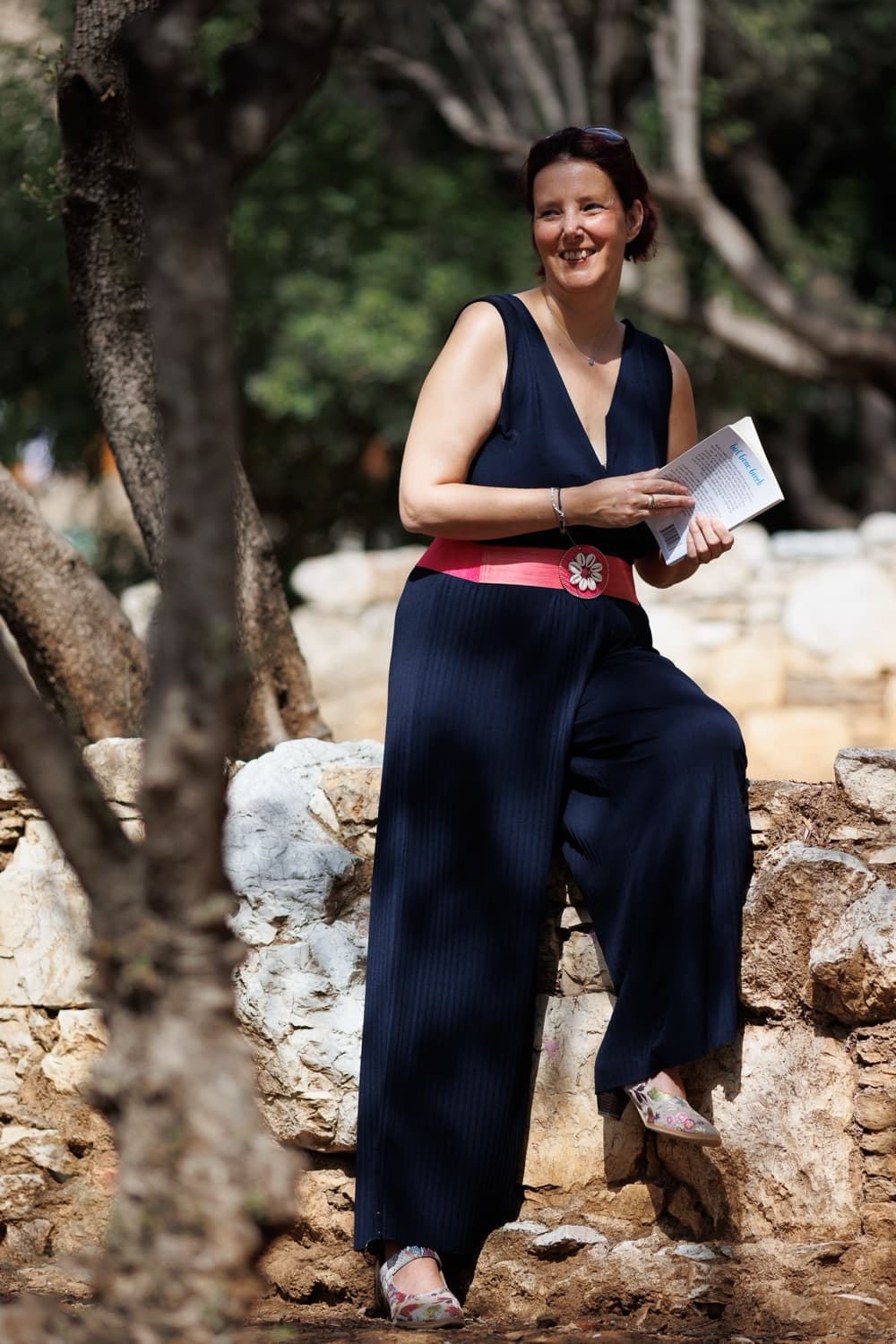 Rebecca Hall sitting on an old stone wall beneath olive trees